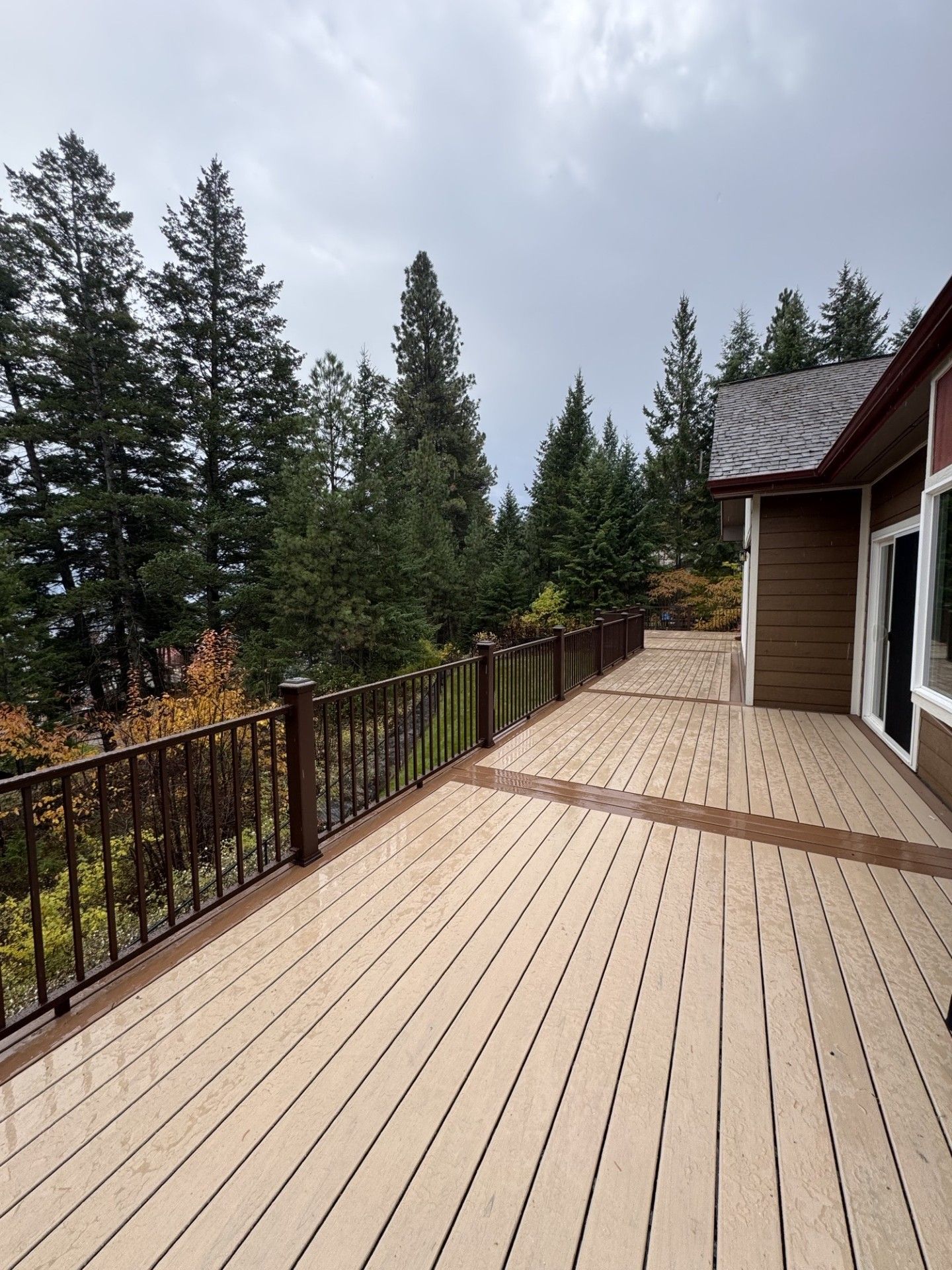 Wooden deck beside a house, with railing, trees, and cloudy sky beyond.