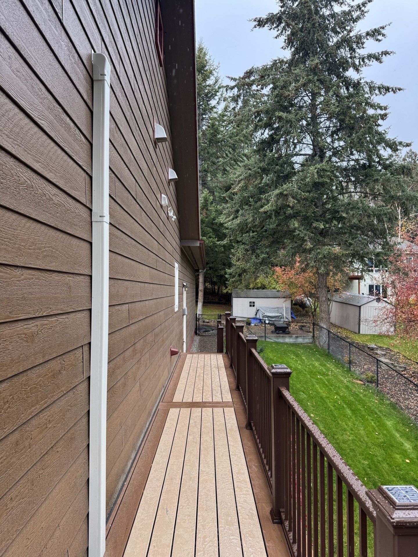 Narrow wooden deck beside a brown house, overlooking a green yard and trees.