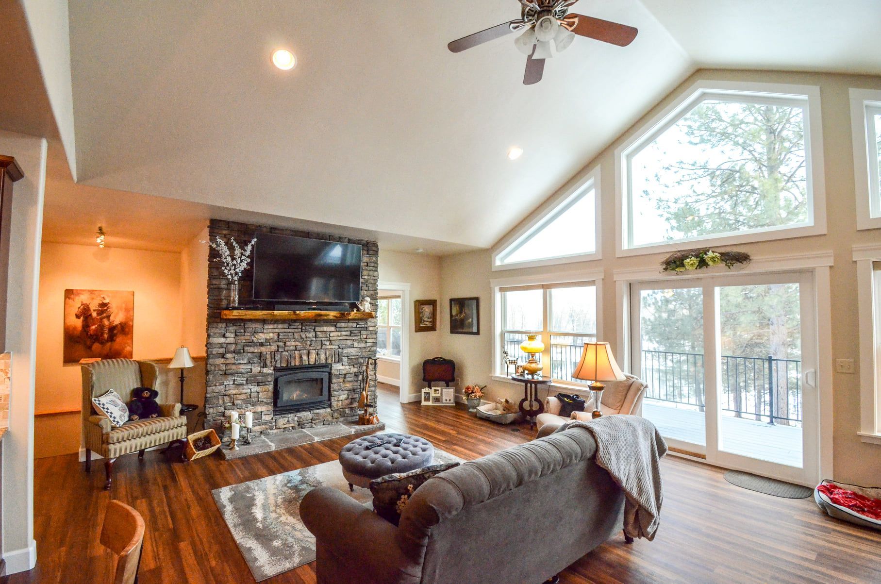 Bright living room with stone fireplace, large windows, gray sofa, hardwood floors, and vaulted ceiling