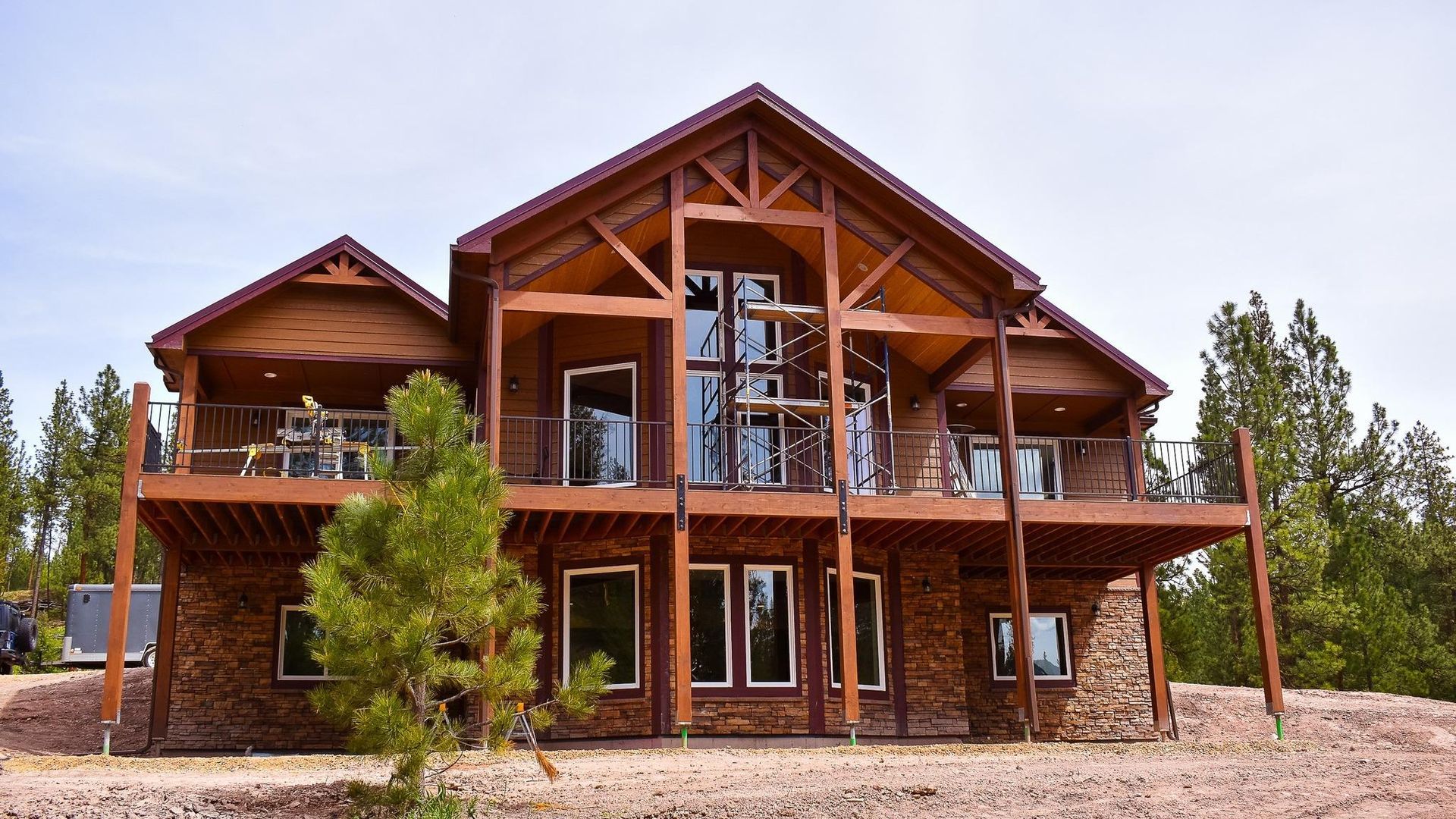 Wooden mountain lodge with a central glass entry and two balconies, surrounded by trees and gravel.