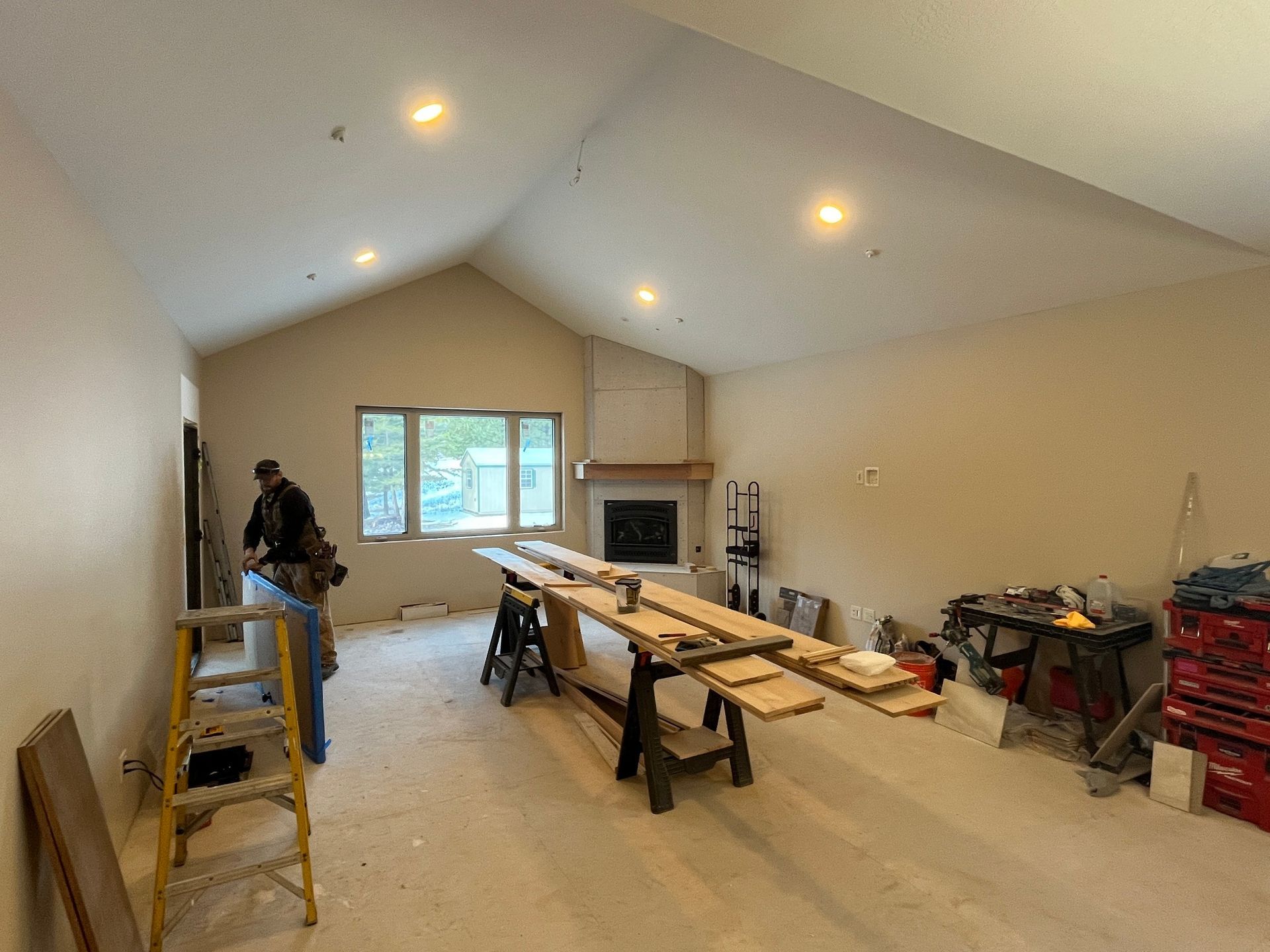 Unfinished living room with workers, sawhorses, lumber, and tools under vaulted ceiling.