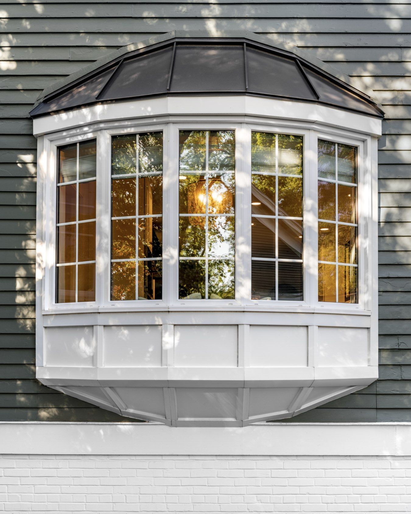 White bay window with black roof on gray-sided house, reflecting sunlight and trees.
