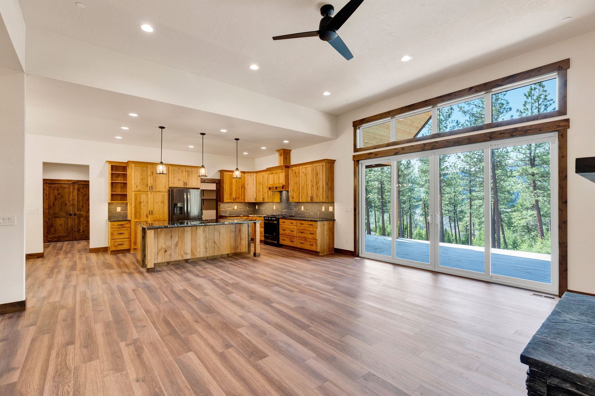 Bright open kitchen and living area with wood floors, vaulted ceiling, large windows, and forest view