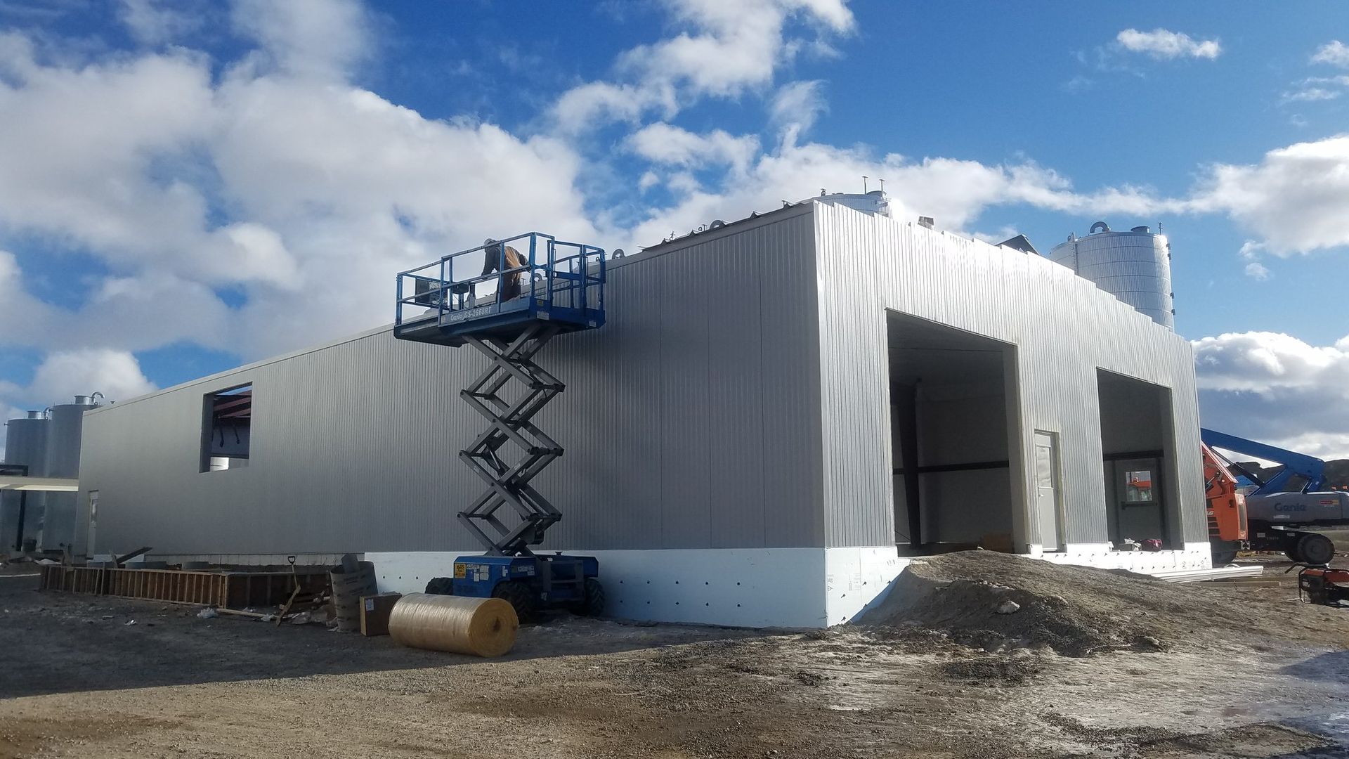 White modular building under construction with a blue scissor lift beside it on a snowy site.