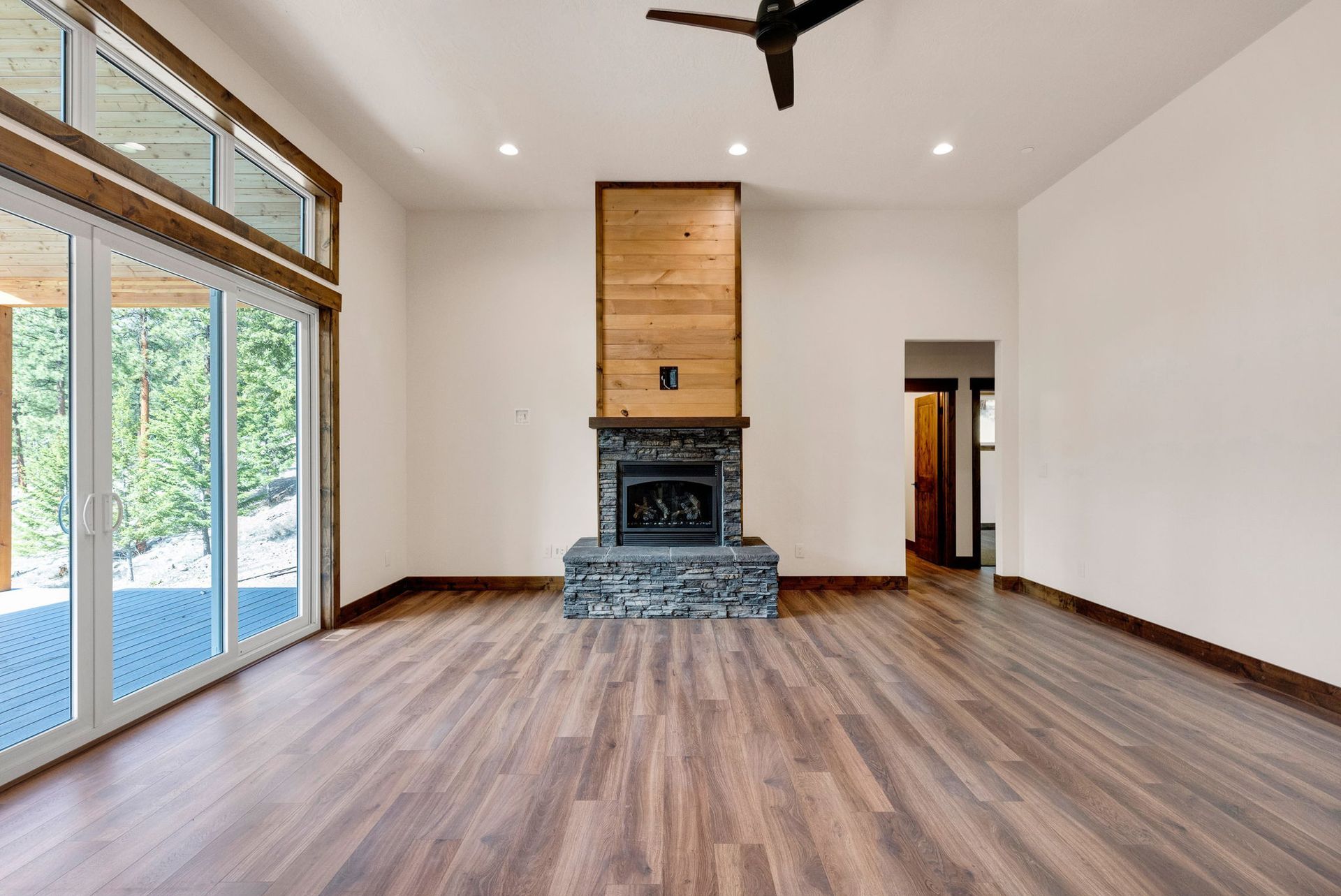 Empty living room with wood floors, large windows, and a central stone fireplace beneath a wood panel.