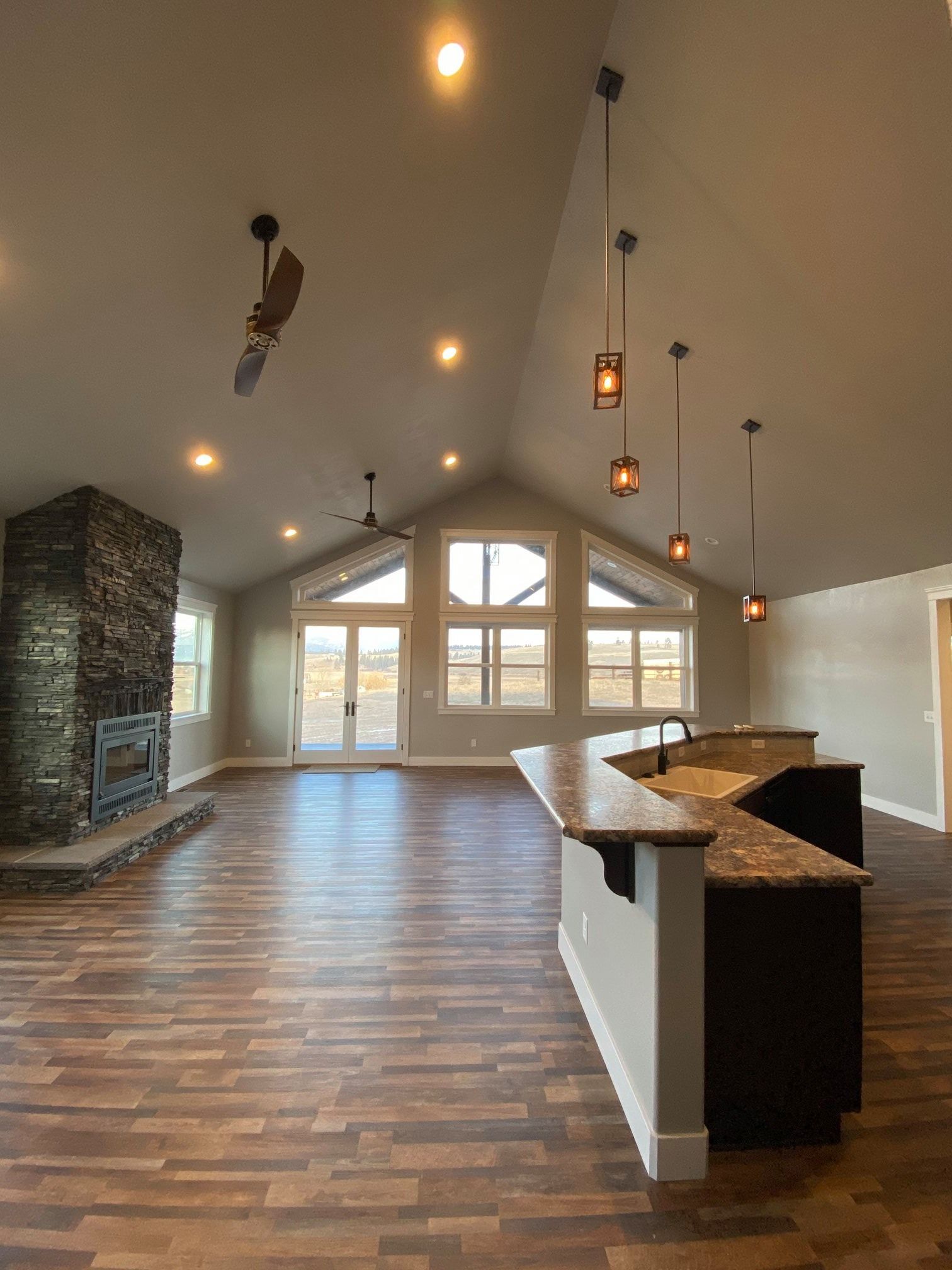 Empty modern living room with vaulted ceiling, stone fireplace, hardwood floors, and large windows.