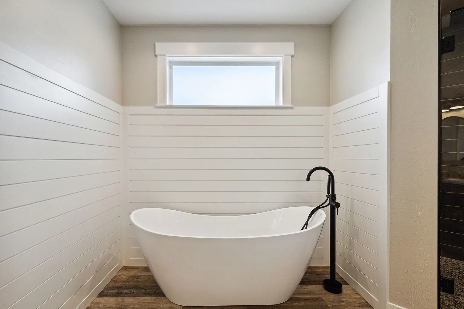 Small bathroom with a white freestanding bathtub, black floor faucet, and window above shiplap walls.