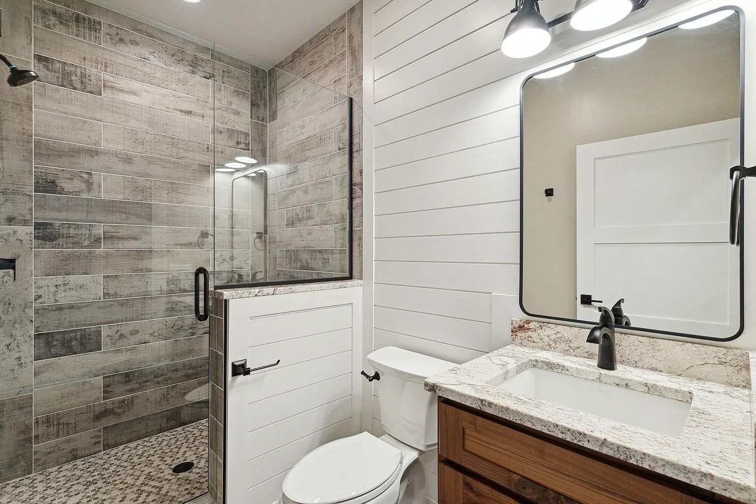 Modern bathroom with tiled shower, glass door, white shiplap walls, and wood vanity with mirror.
