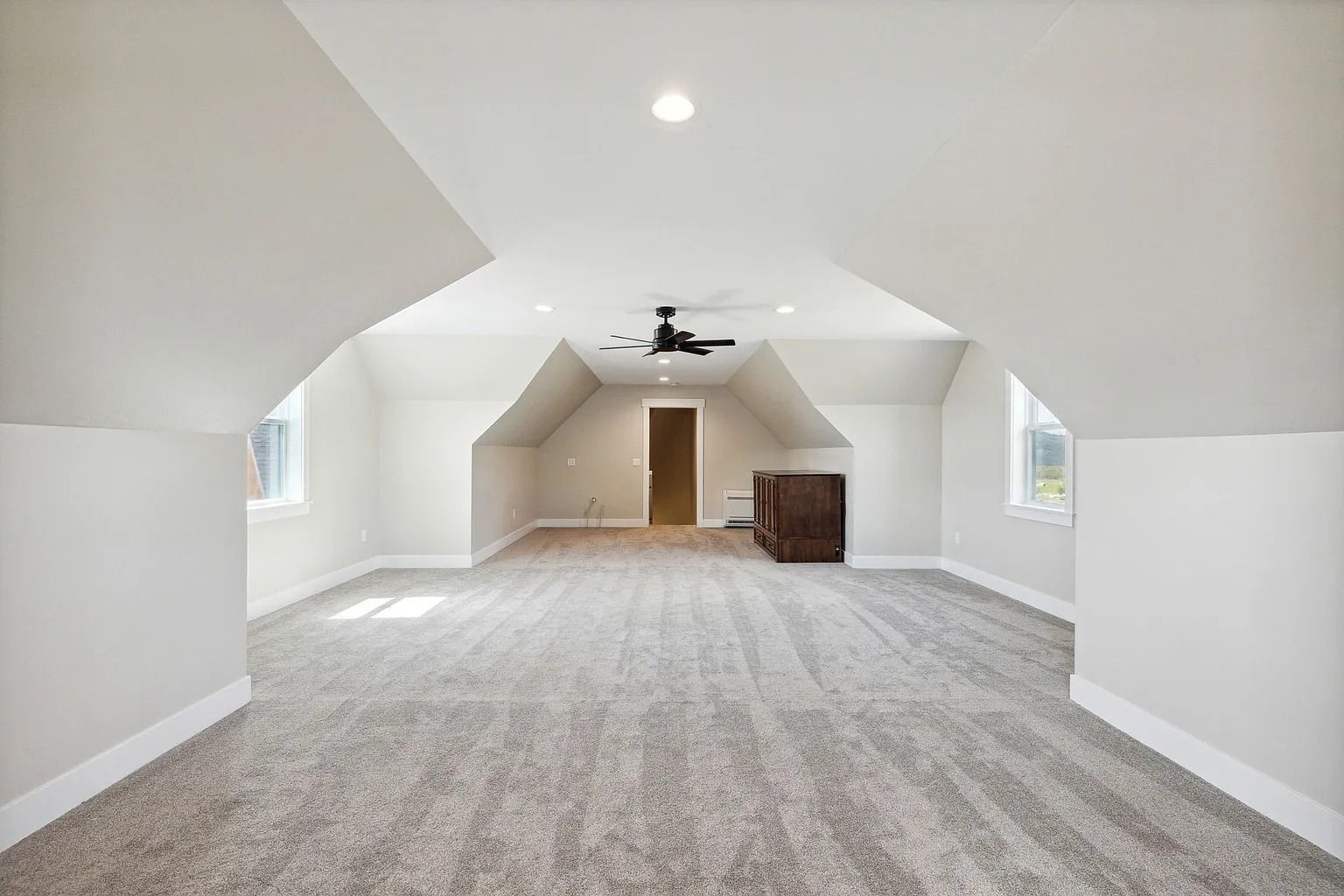Empty carpeted attic room with white walls, sloped ceiling, ceiling fan, and wood dresser at the far end
