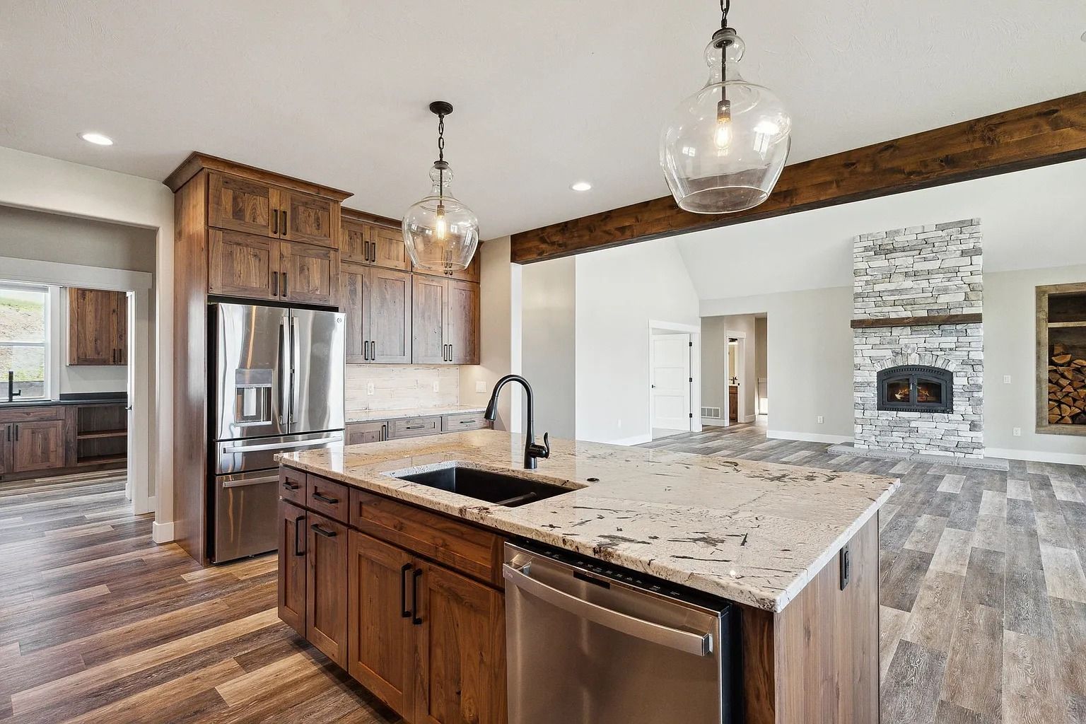 Bright open kitchen with granite island, stainless appliances, pendant lights, and living room with stone fireplace