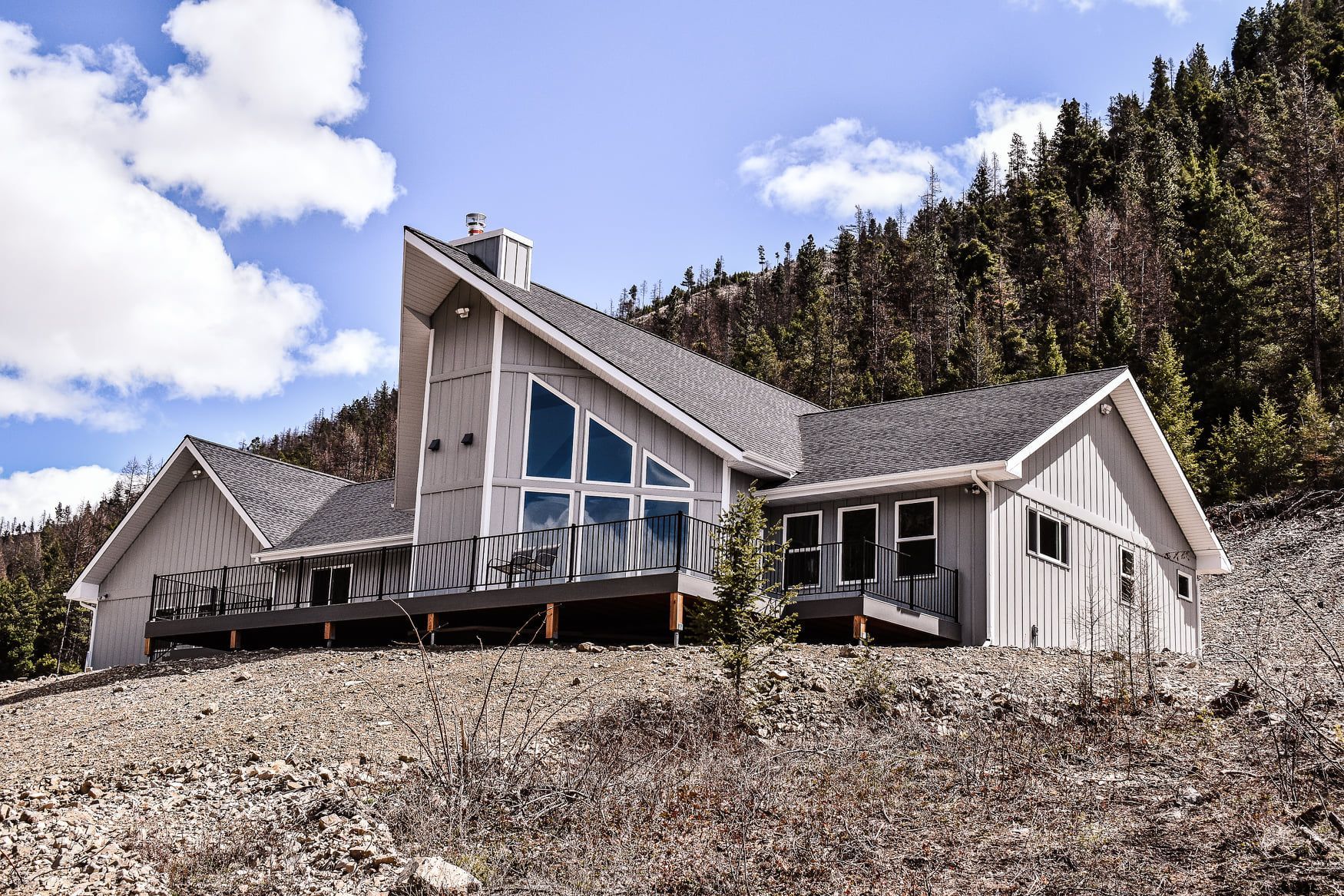 A modern cabin with large windows sits on a rocky hillside under a blue sky with clouds.