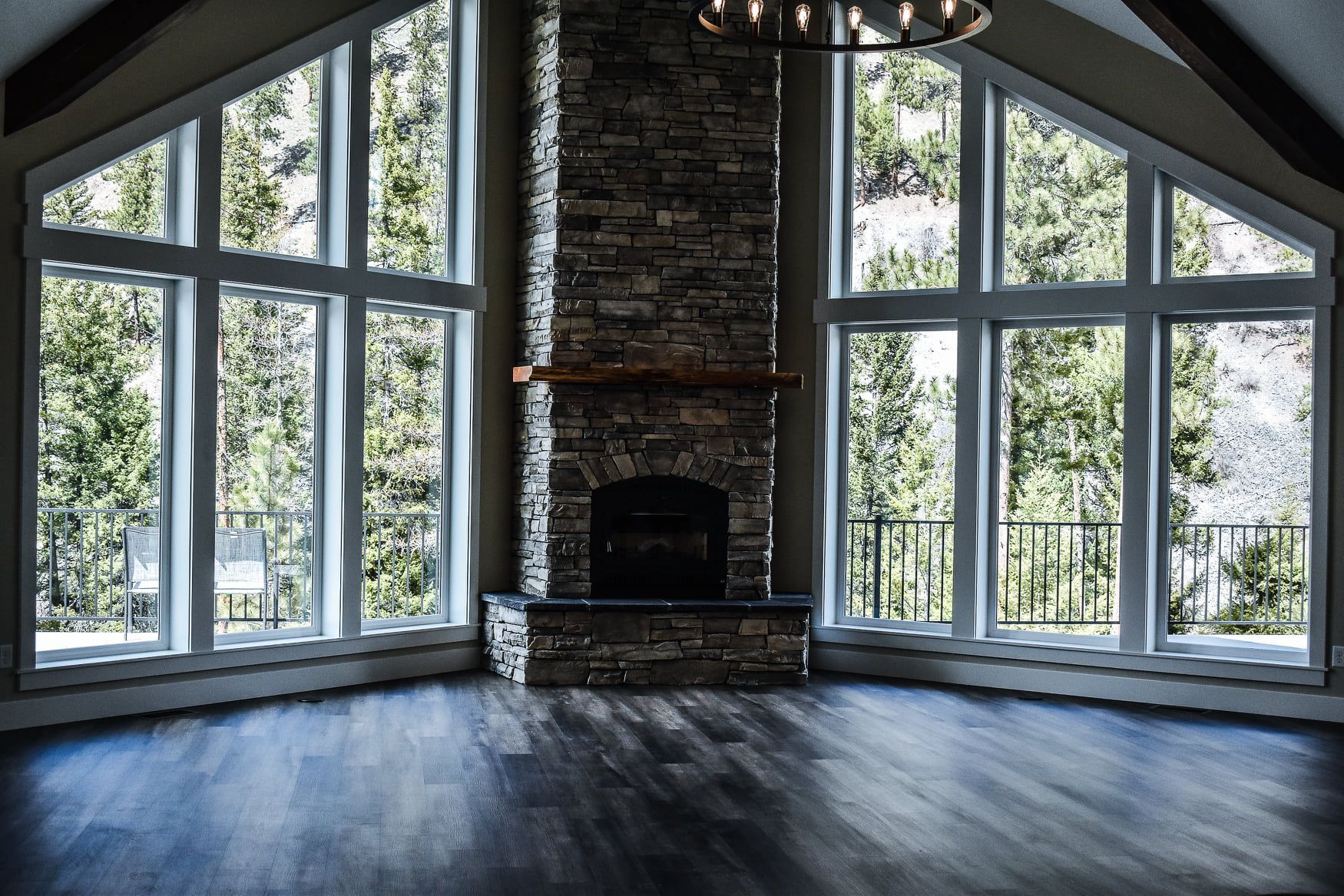 Empty room with a stone fireplace, vaulted windows, and dark wood floors overlooking trees.