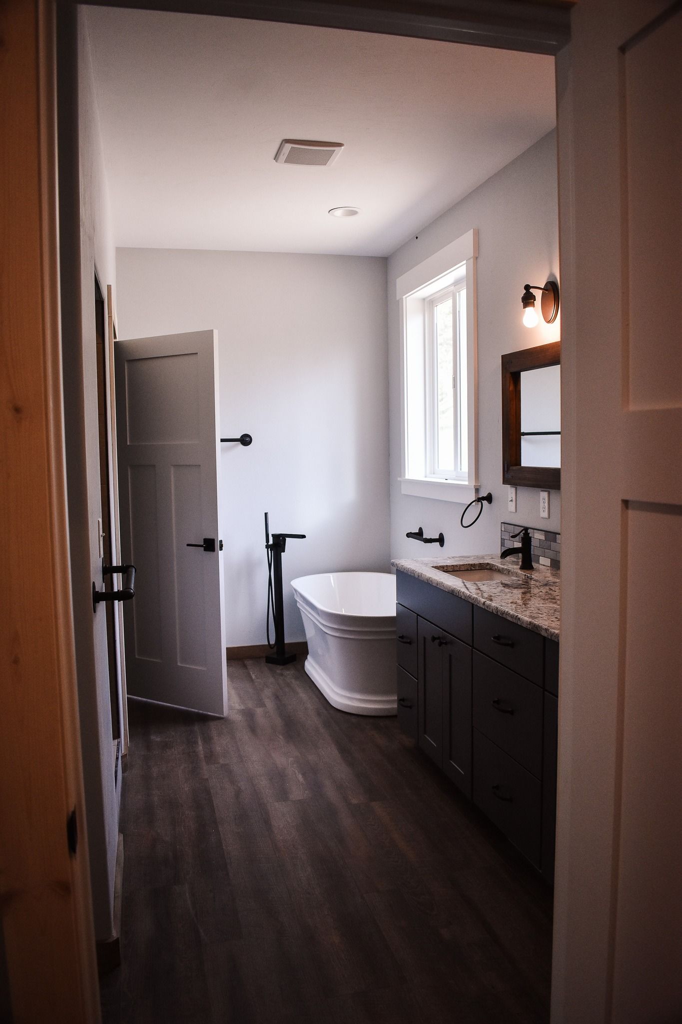 Modern bathroom with white soaking tub, dark vanity, window, and open door in a narrow hallway