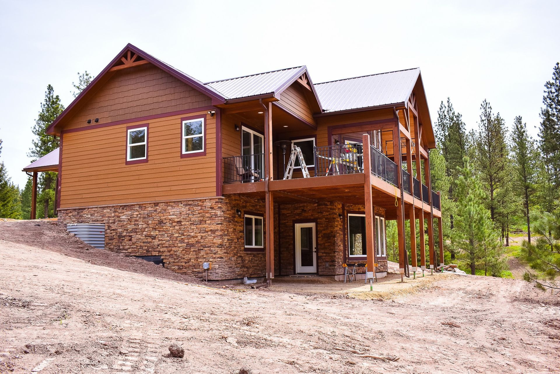 Rustic two-story wooden cabin with stone foundation on a dirt lot surrounded by pine trees.
