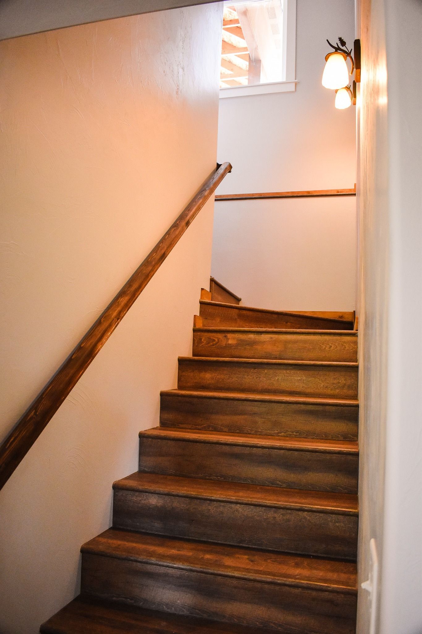Narrow wooden staircase with white walls and a wall sconce light at the landing.