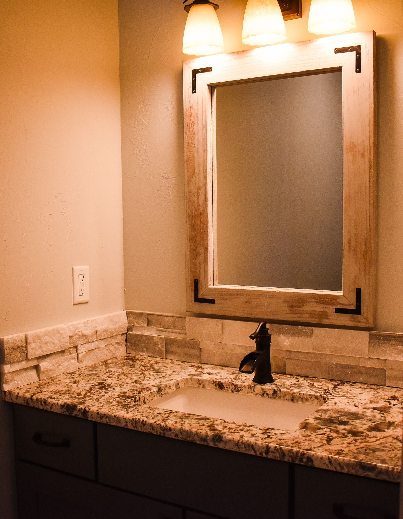 Bathroom vanity with granite countertop, stone backsplash, and lit mirror above the sink