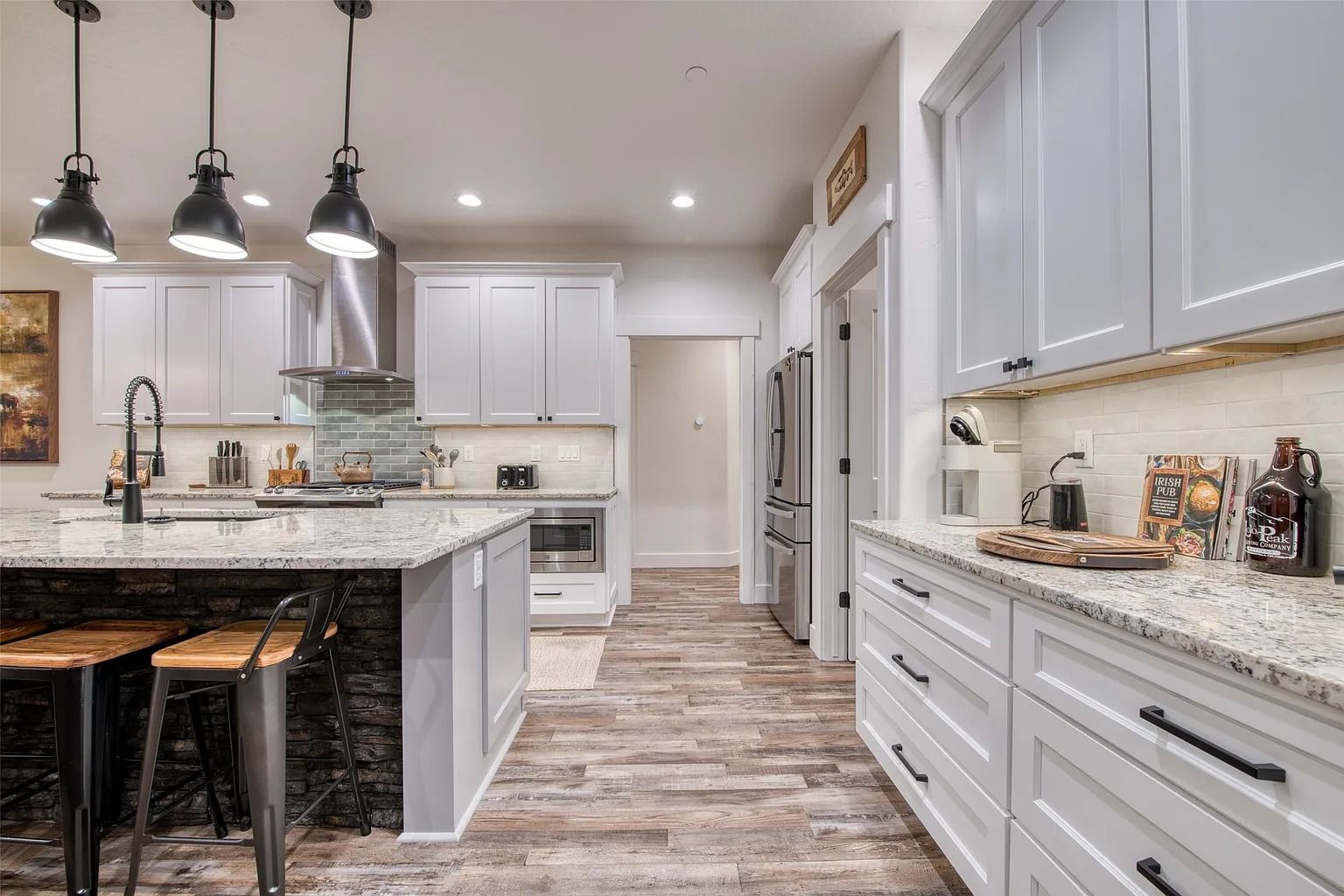 Modern white kitchen with marble counters, dark pendant lights, and a rustic wood floor.