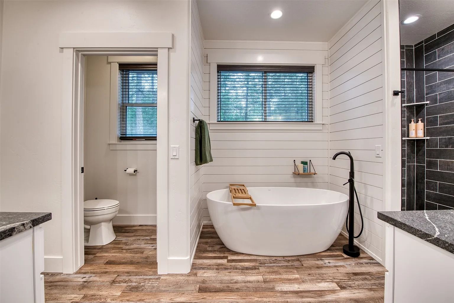 Modern bathroom with freestanding tub, toilet, vanity, and two windows with blinds
