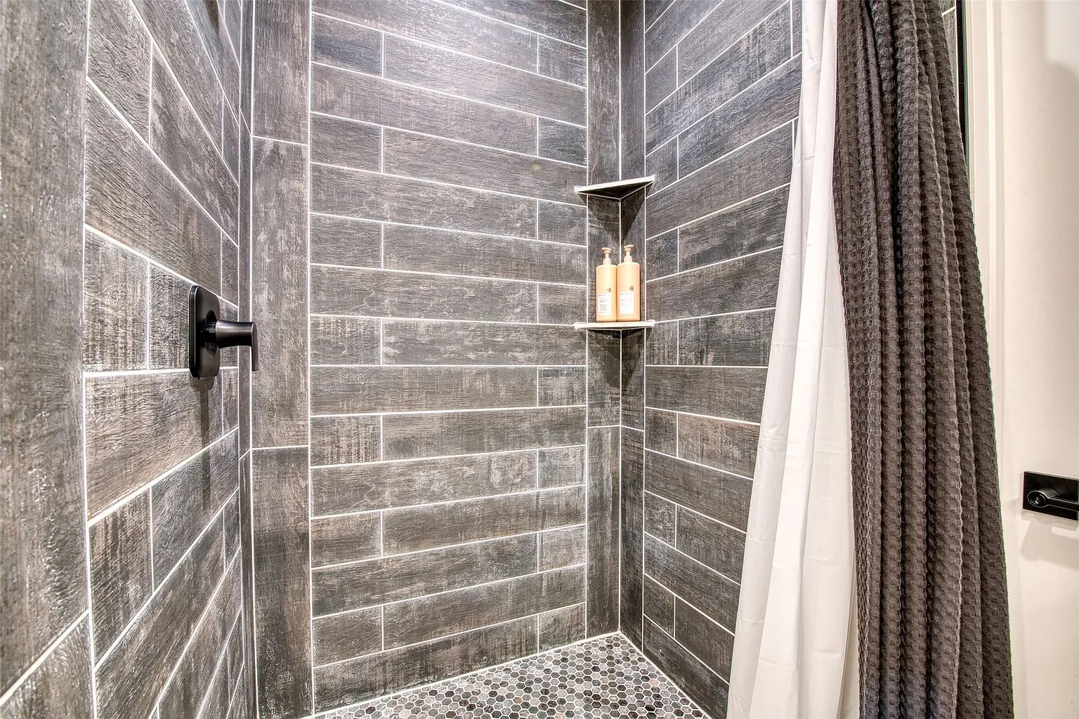 Modern gray tiled shower with a black fixture, small corner shelf, and striped curtain