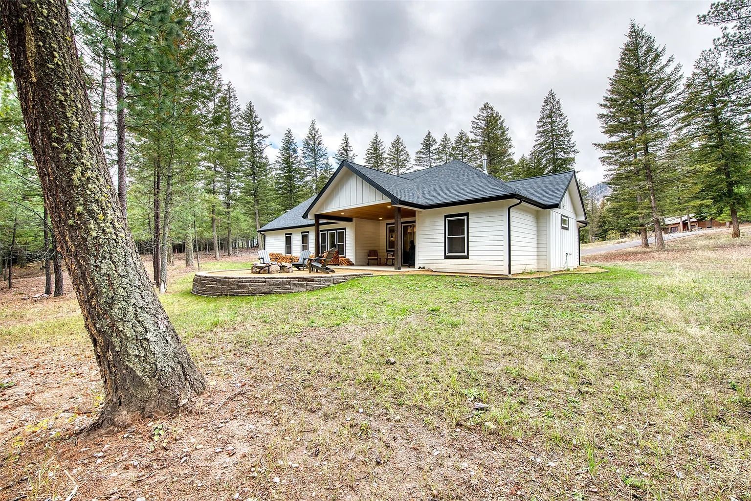 White house with porch in a wooded yard, surrounded by tall pine trees and a grassy clearing.
