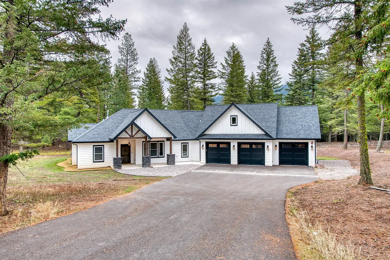 Single-story gray house with blue roof and three-car garage, surrounded by trees and a gravel driveway.