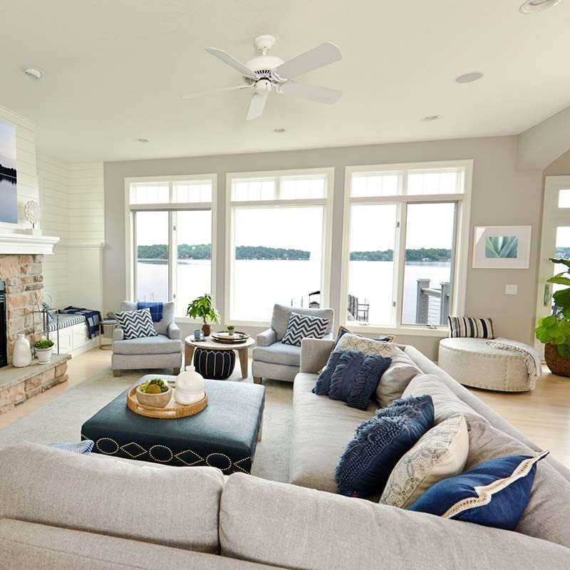 Bright lakefront living room with gray sofas, large windows, white ceiling fan, and beige decor