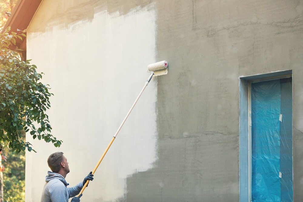 A Man is Painting the Side of a House With a Paint Roller — Prota NT in Howard Springs, NT