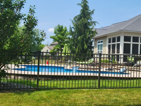 A fence surrounds a large swimming pool in the backyard of a house.