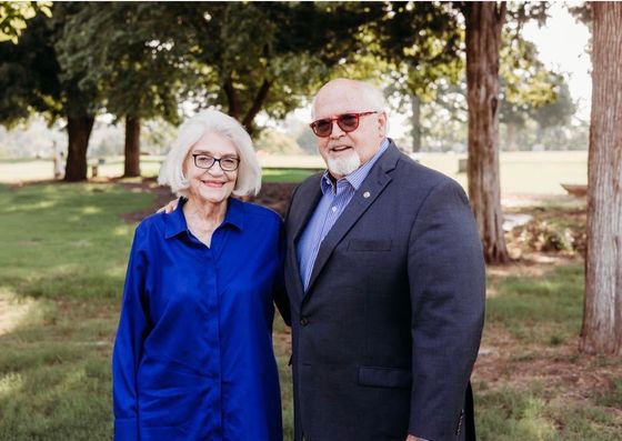 Smiling older couple outdoors; man in blue shirt standing behind woman in white shirt, with trees in the background