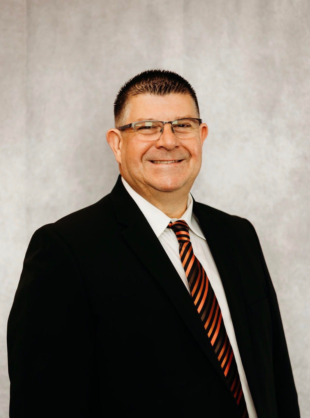 Smiling man in navy suit and patterned tie standing before a wooden wall