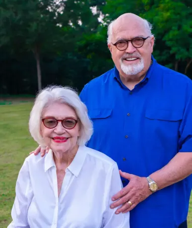 Smiling older couple outdoors; man in blue shirt standing behind woman in white shirt, with trees in the background
