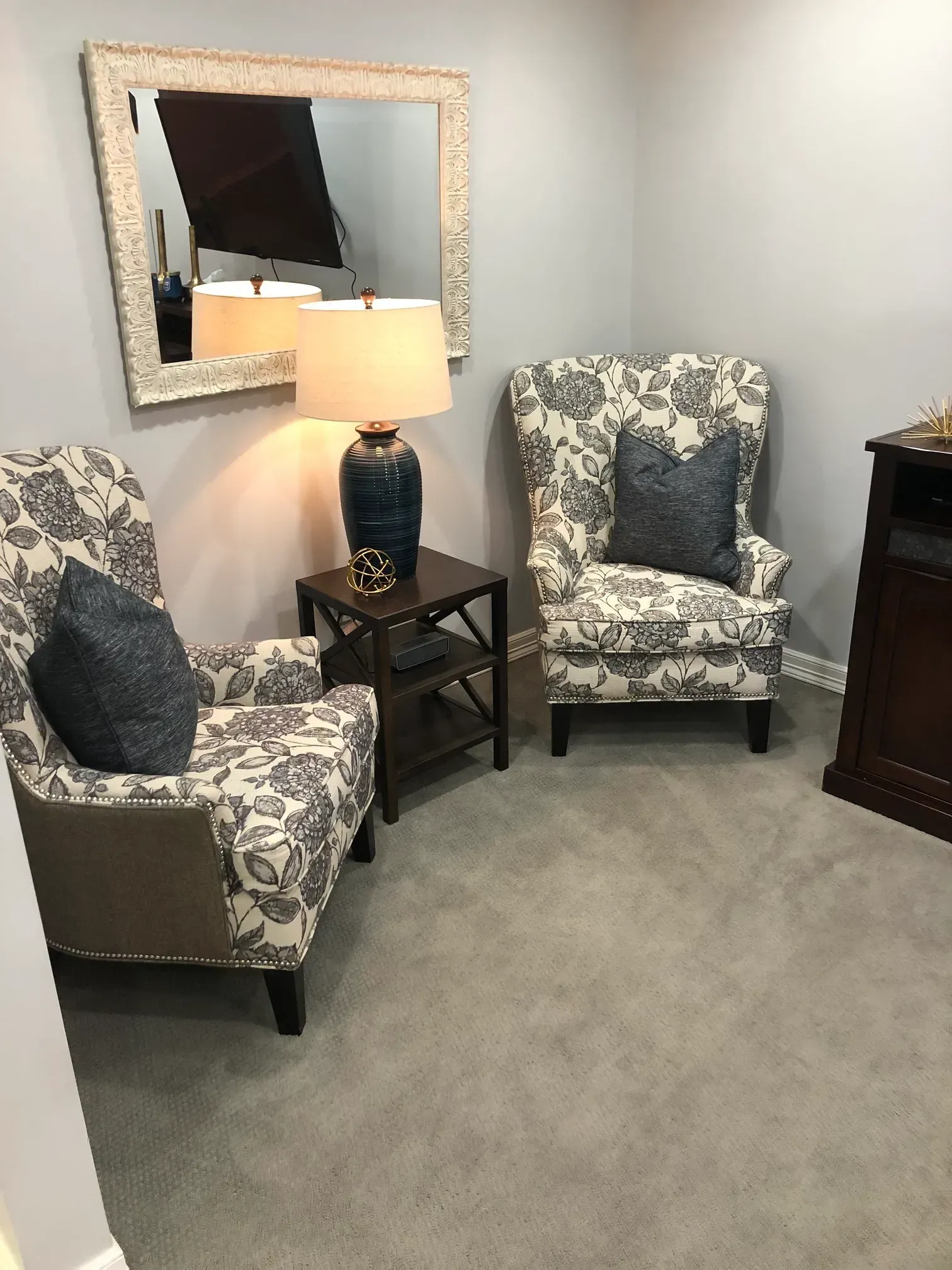 Cozy sitting area with two patterned chairs, lamp table, wall mirror, and gray carpet