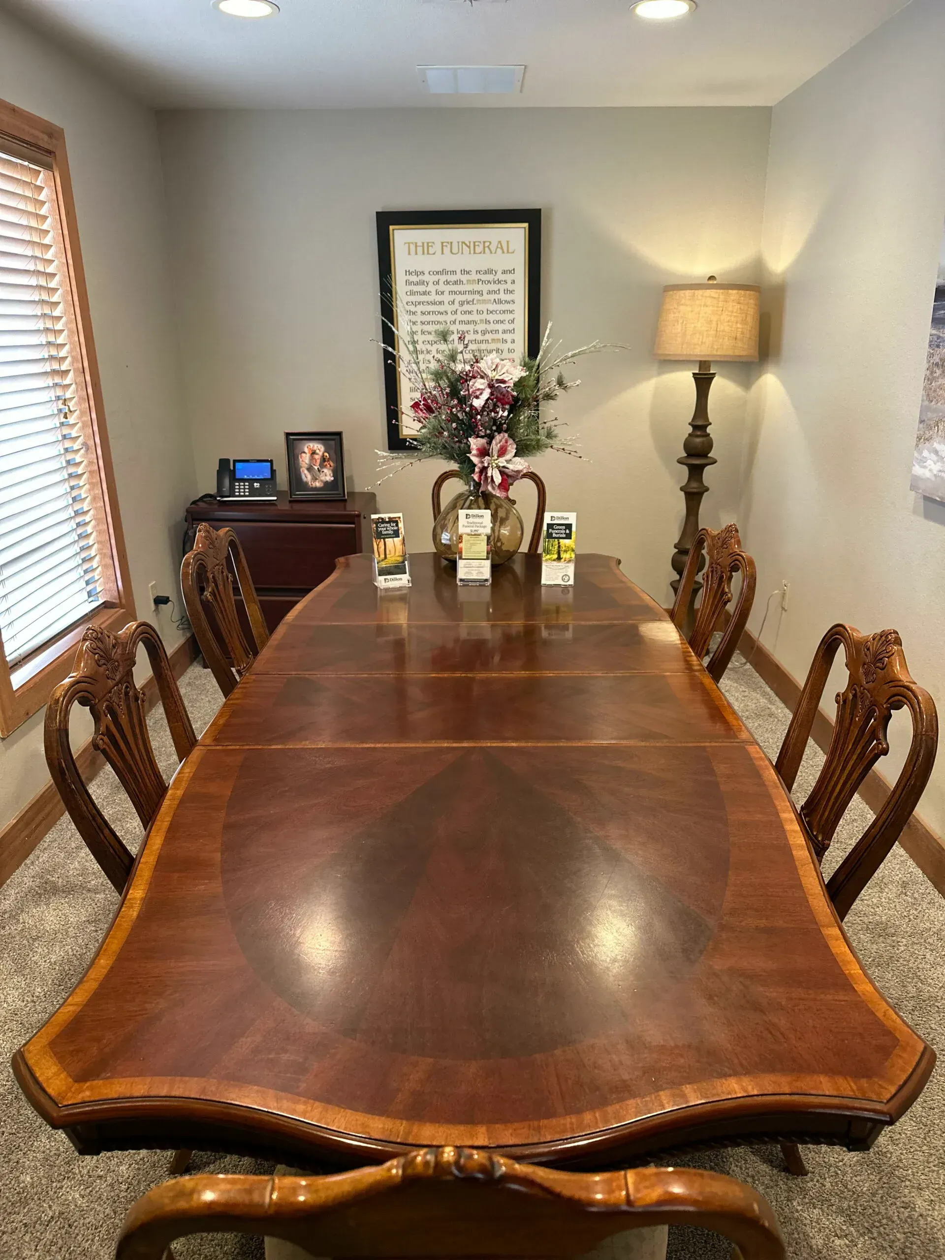 Dining room with a wooden table, chairs, lamp, and flowers against a light gray wall.