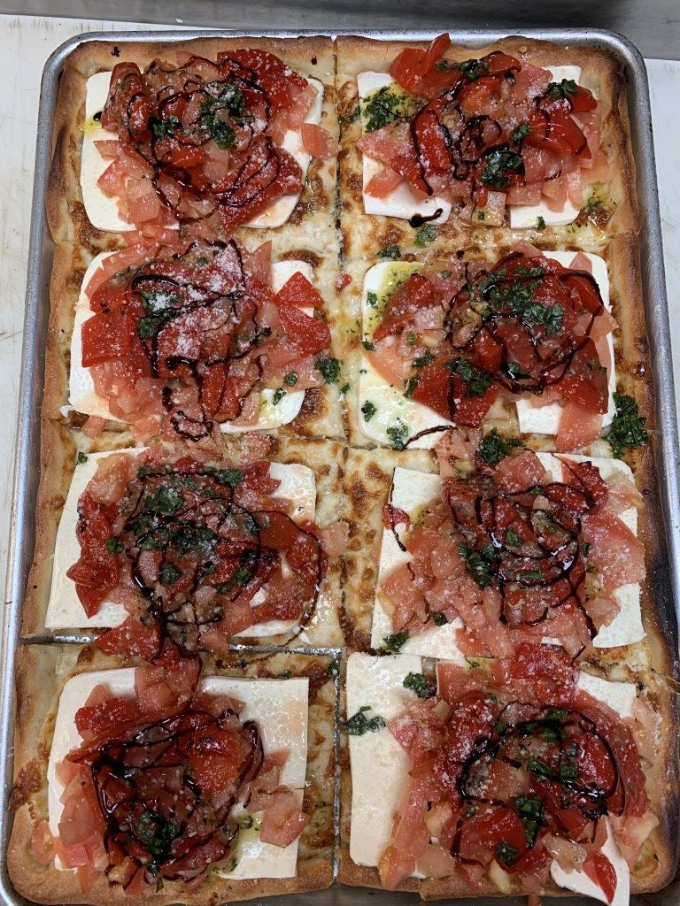 Bruschetta appetizer on a rectangular tray. Six squares of bread topped with tomatoes, cheese, herbs, and balsamic glaze.