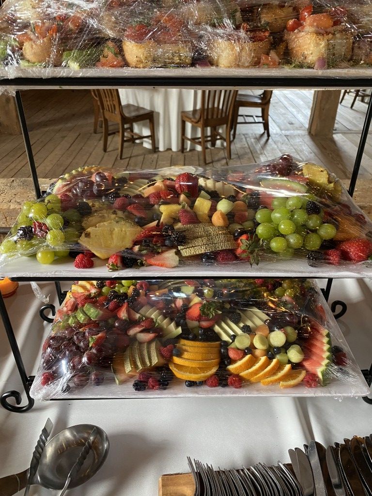 Three-tiered display of various fruits on white platters, covered in plastic wrap. Silverware on table.