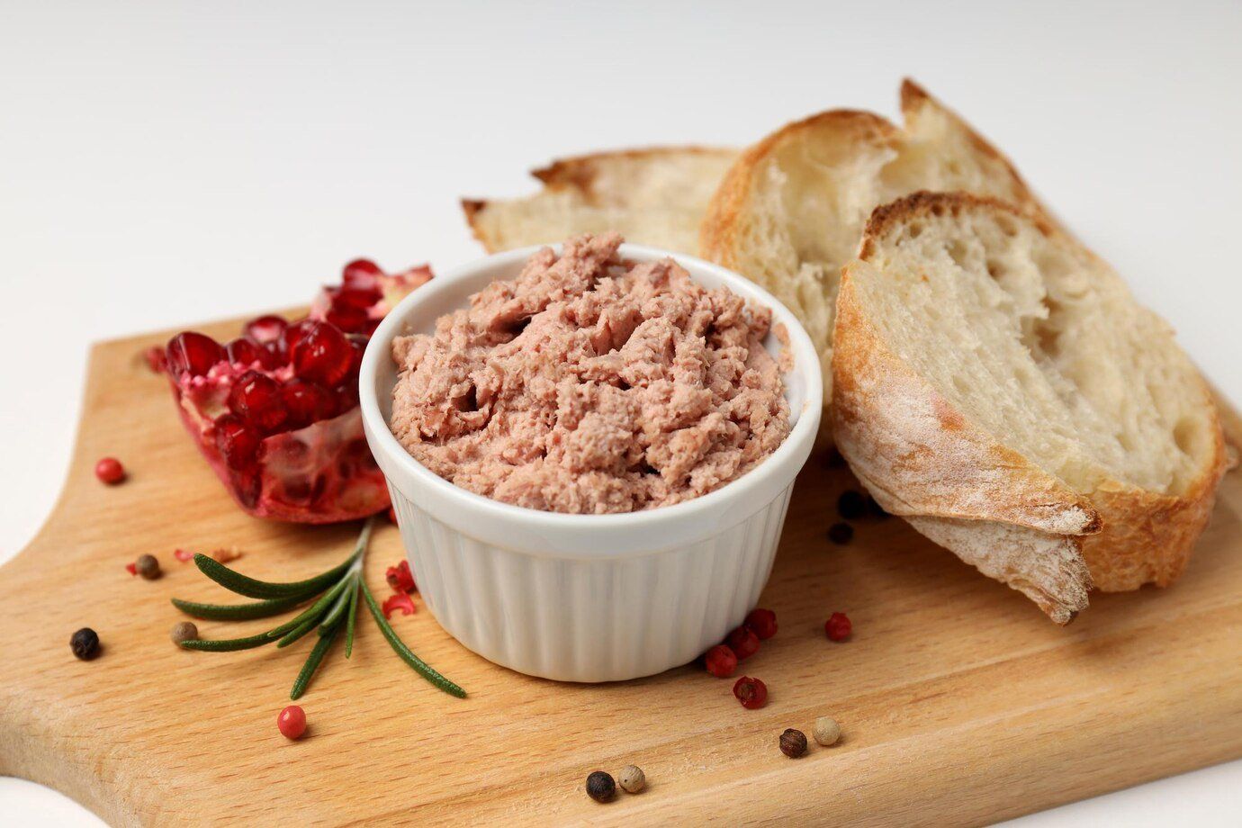 Pâté in white bowl with sliced bread, pomegranate, rosemary, and peppercorns on a wooden board.