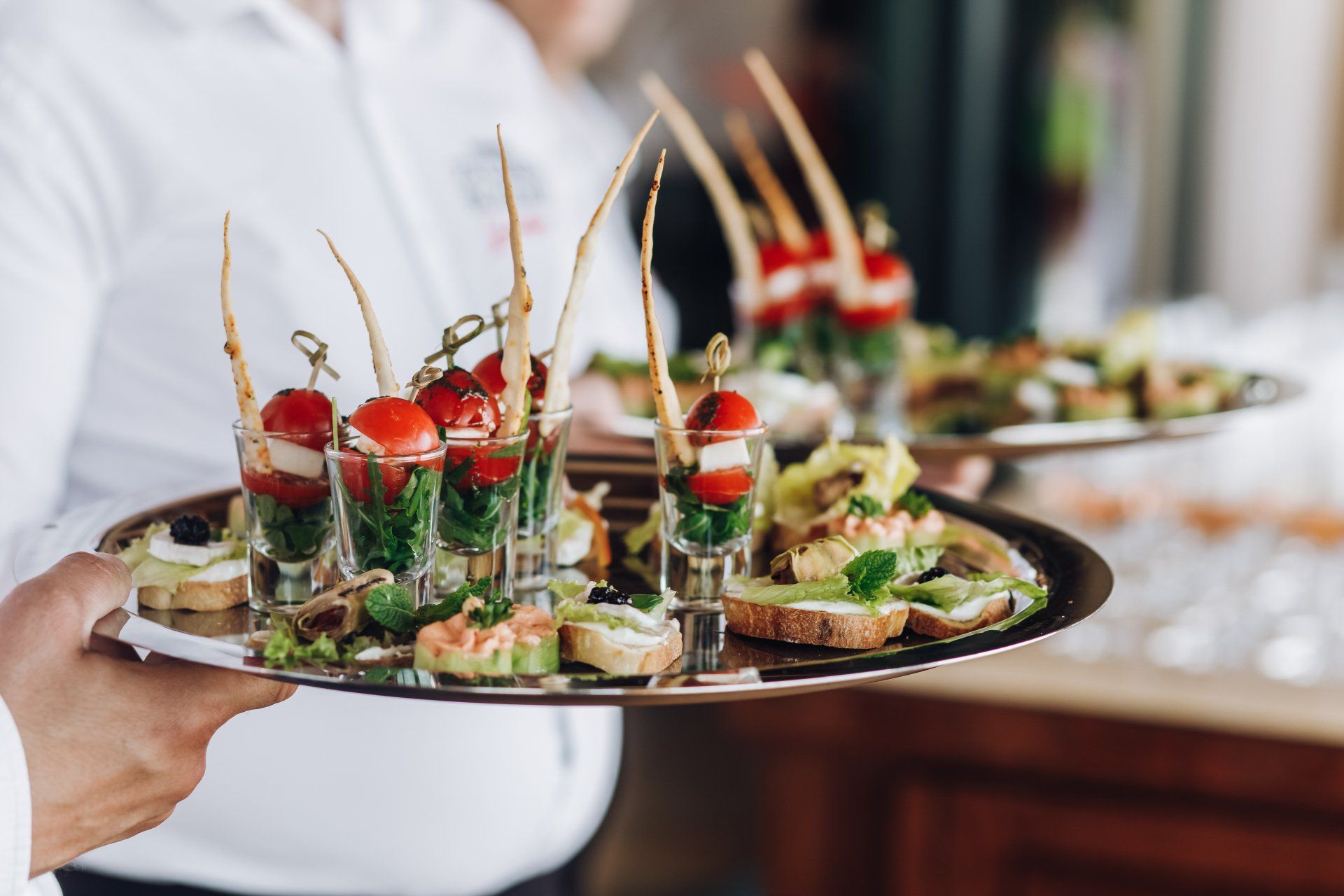 Waiter holding a tray of appetizers, including shot glasses with tomatoes, and open-faced sandwiches.