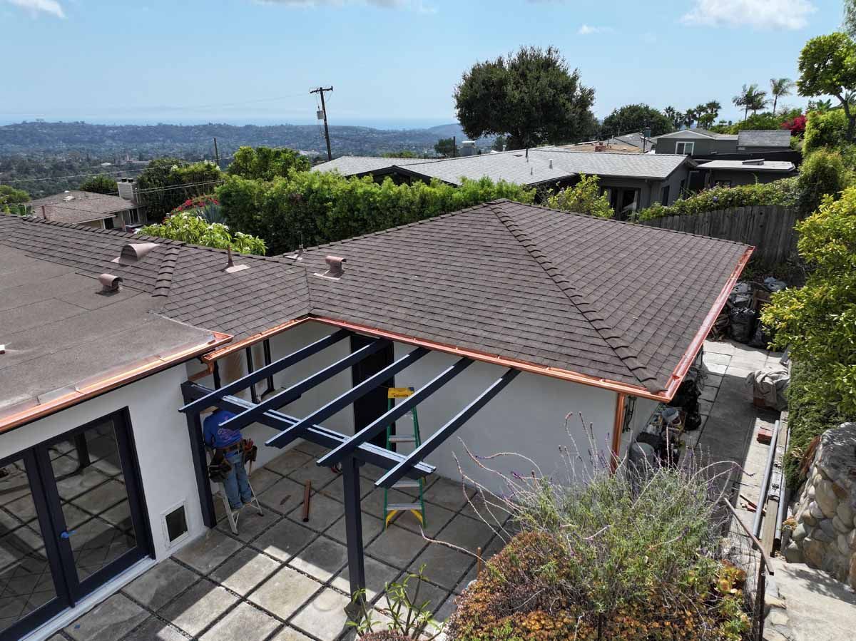 Rooftop with brown shingles and metal beams overlooking a suburban neighborhood and distant water.