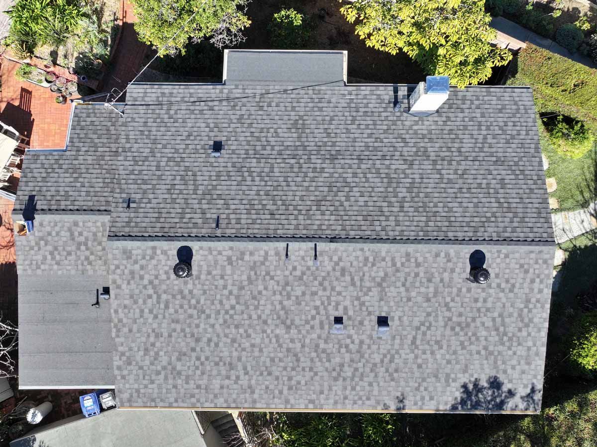 Aerial view of a gray shingled house roof with vents, skylight, and surrounding trees.