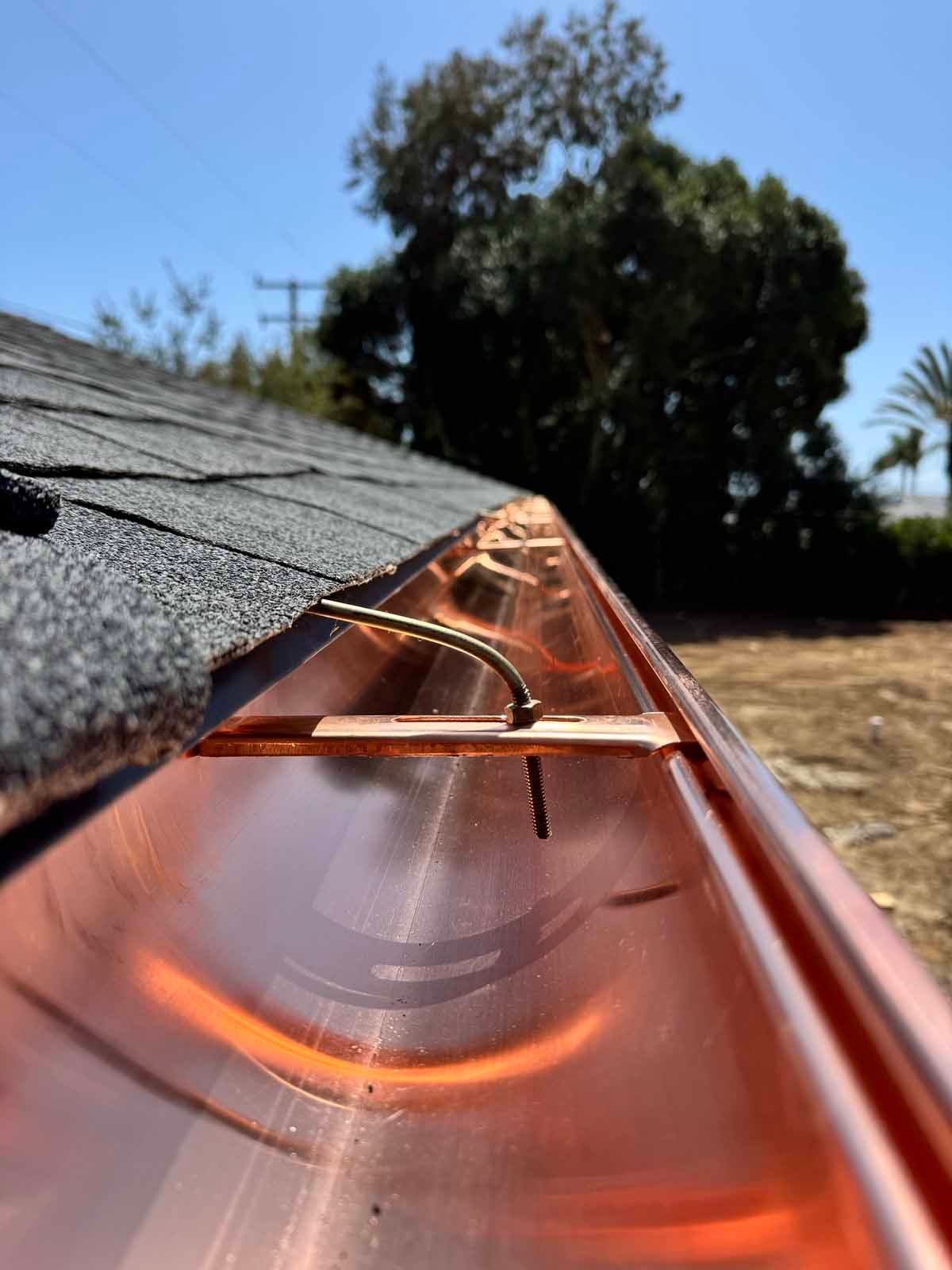 Copper gutter on shingled roof with clear water flowing, outdoors under blue sky