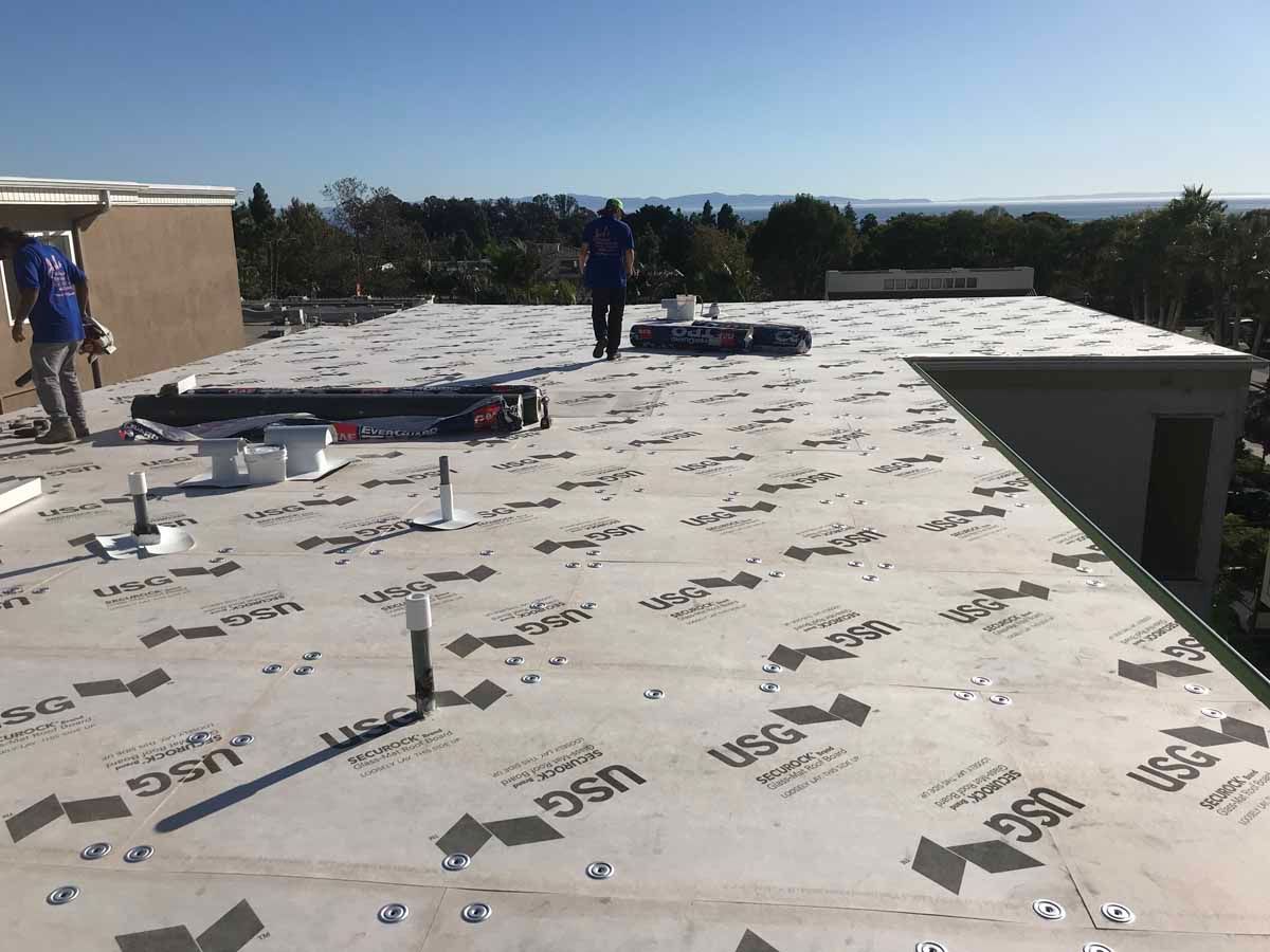 Rooftop under construction with workers, white roofing membrane, tools, and a clear sky in the background