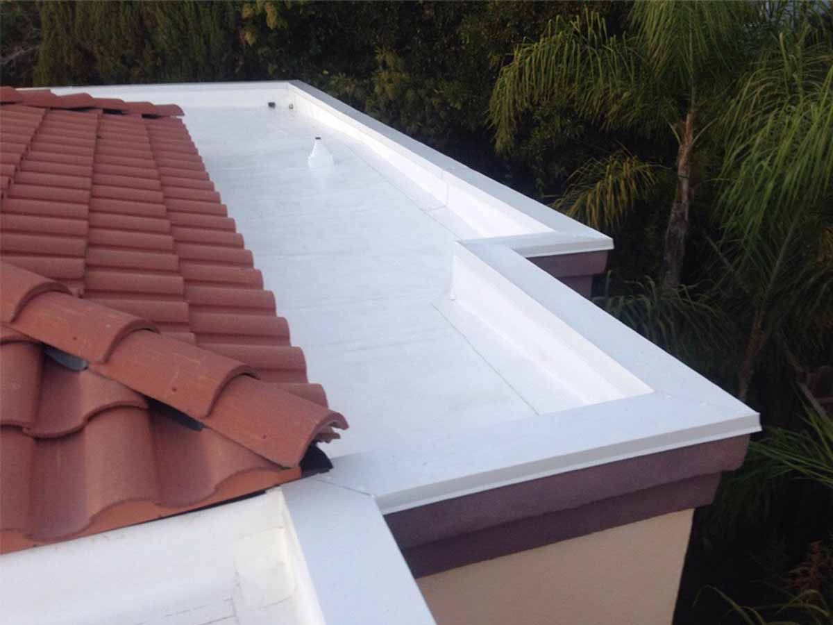 Flat roof with white waterproofing beside red roof tiles and trees in the background