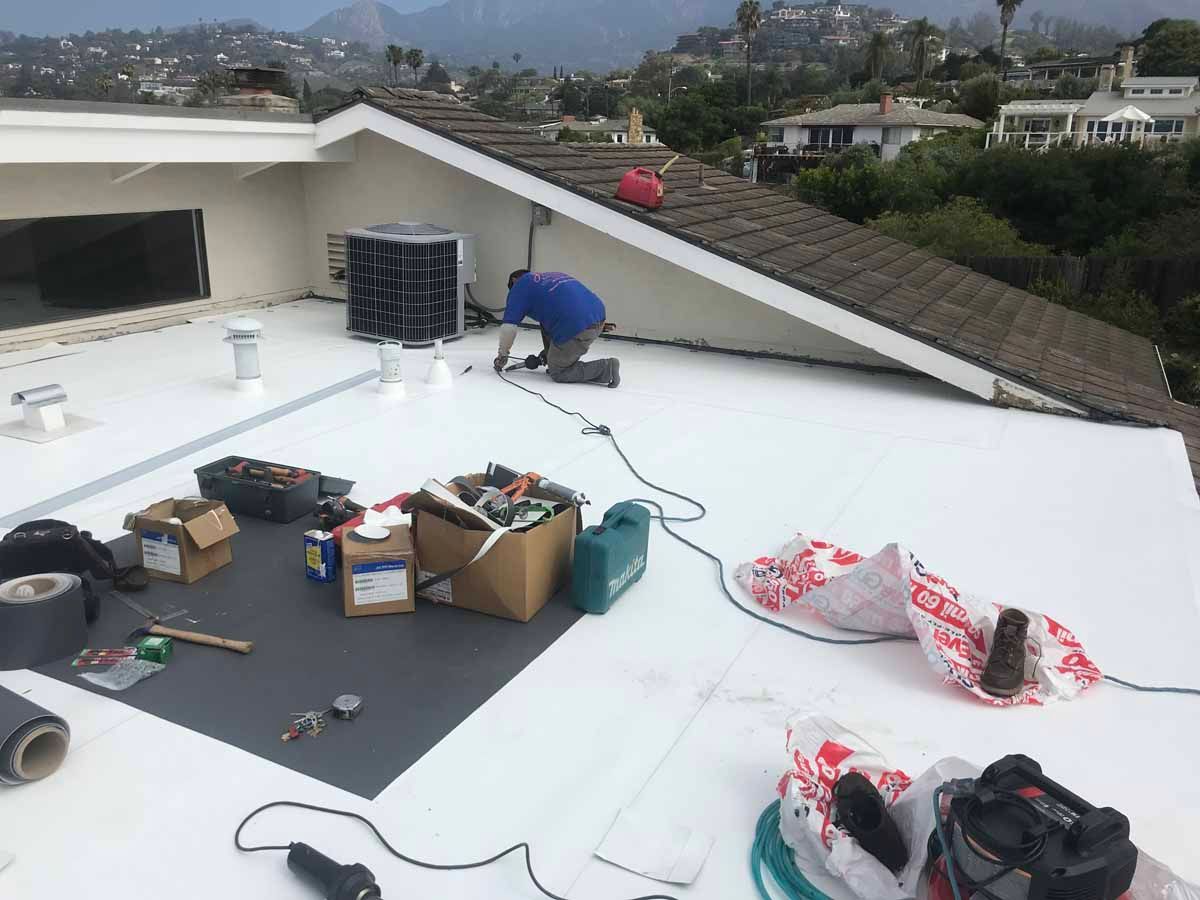 Worker on a white rooftop arranging tools and materials near a sloped roof under repair.