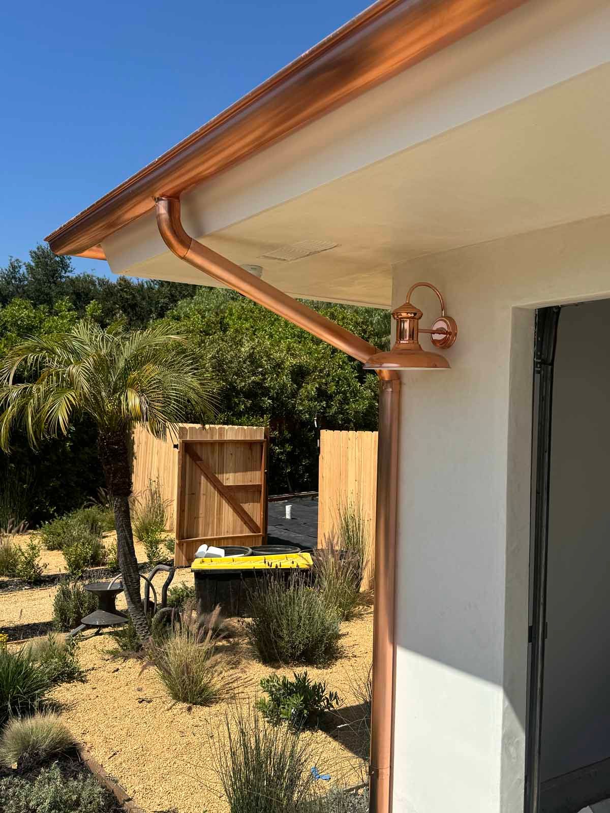 Side of a white house with a copper rain gutter, red outdoor light, and a backyard gate and grill beyond the lawn
