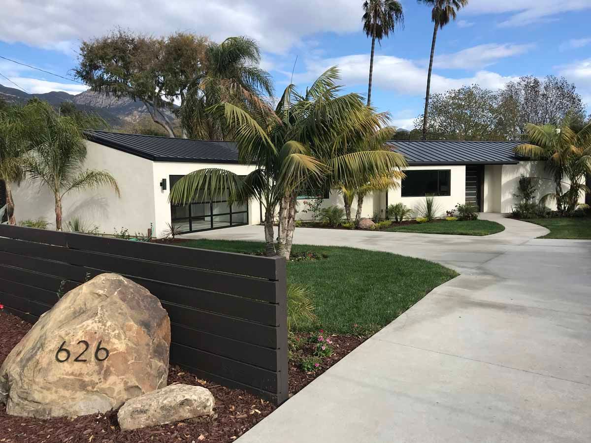 Modern white house with black roof, palm trees, and driveway beside a stone marked 626.
