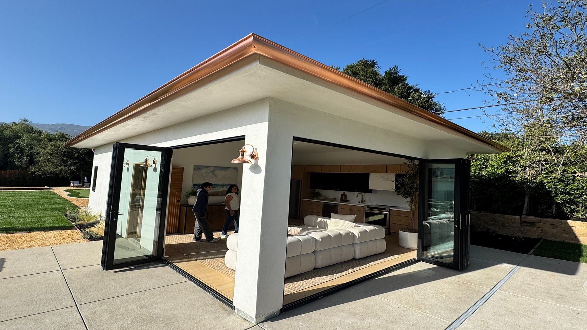 Open-air modern lounge pavilion with white sofas and glass doors in a sunny outdoor setting