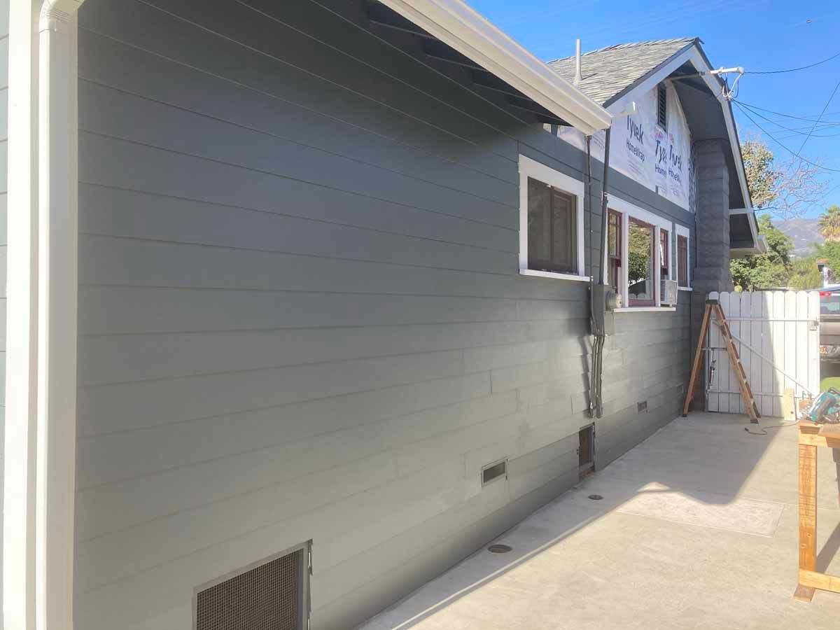 Side view of a gray house exterior with windows, a concrete walkway, and a fenced yard.