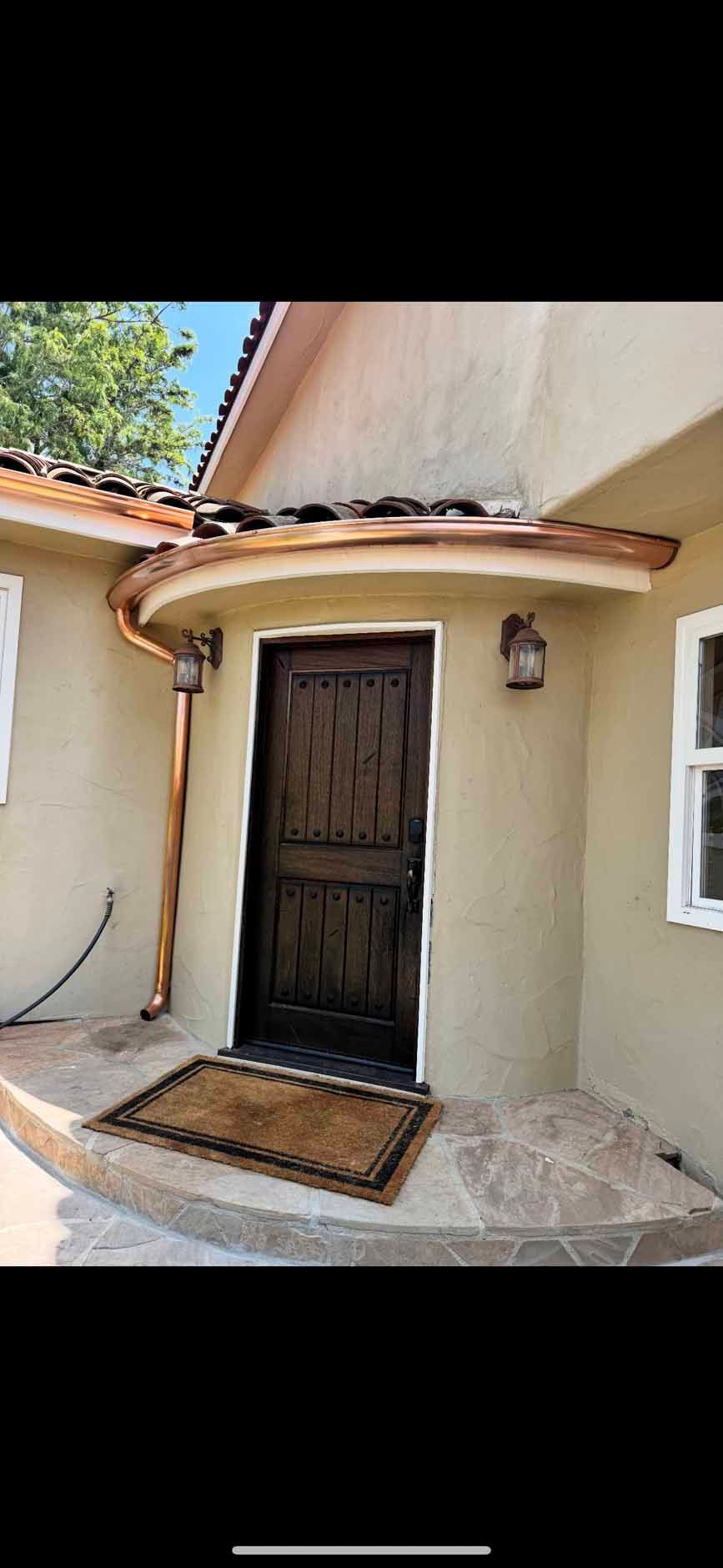 Front entrance of a beige house with a dark wooden door, small porch, and brown doormat under a tiled roof.