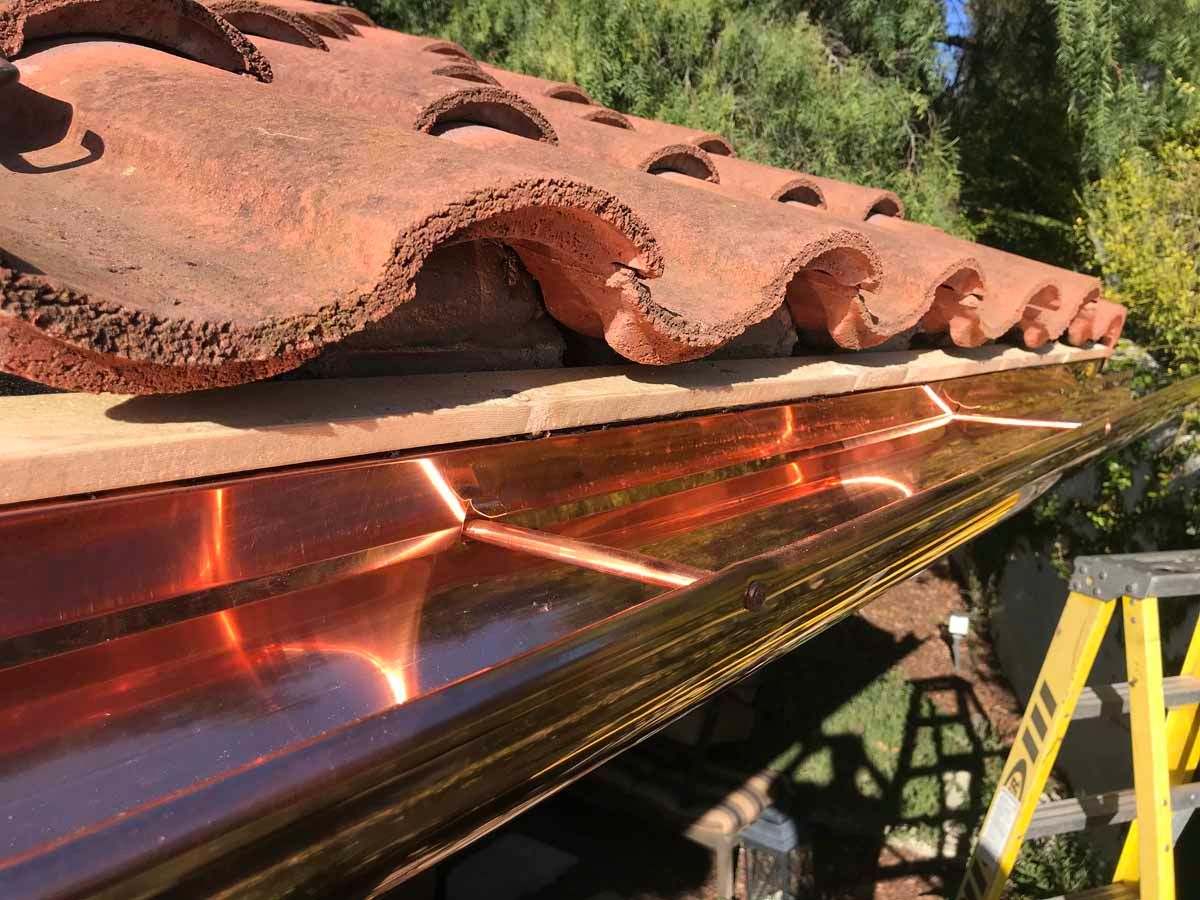 Copper gutter installed beneath a red clay tile roof, with a yellow ladder in the background