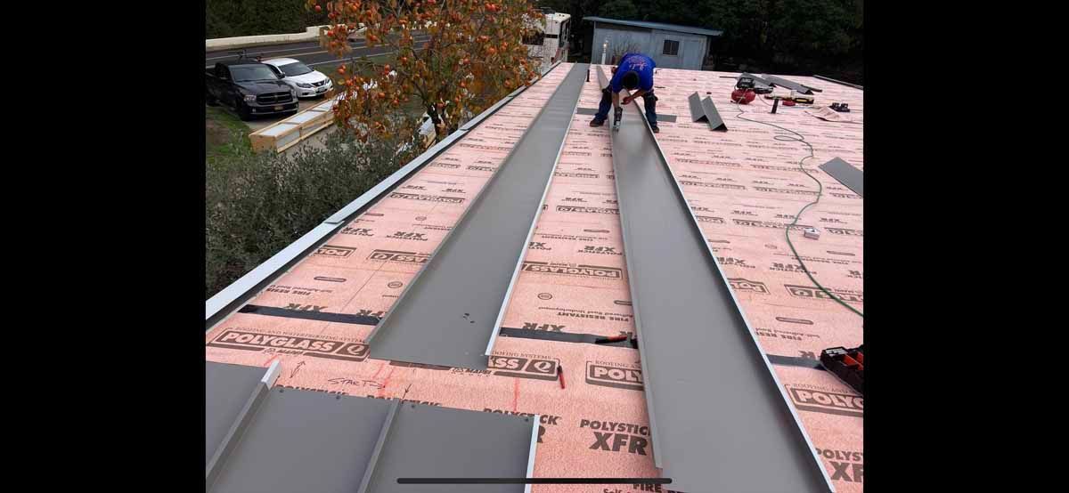 Workers installing long metal roof panels on a pink roof, with a car visible at left.
